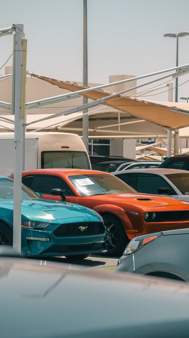 Cars parked beneath shade canopies in a sunlit outdoor lot, including an orange sports car and teal car.