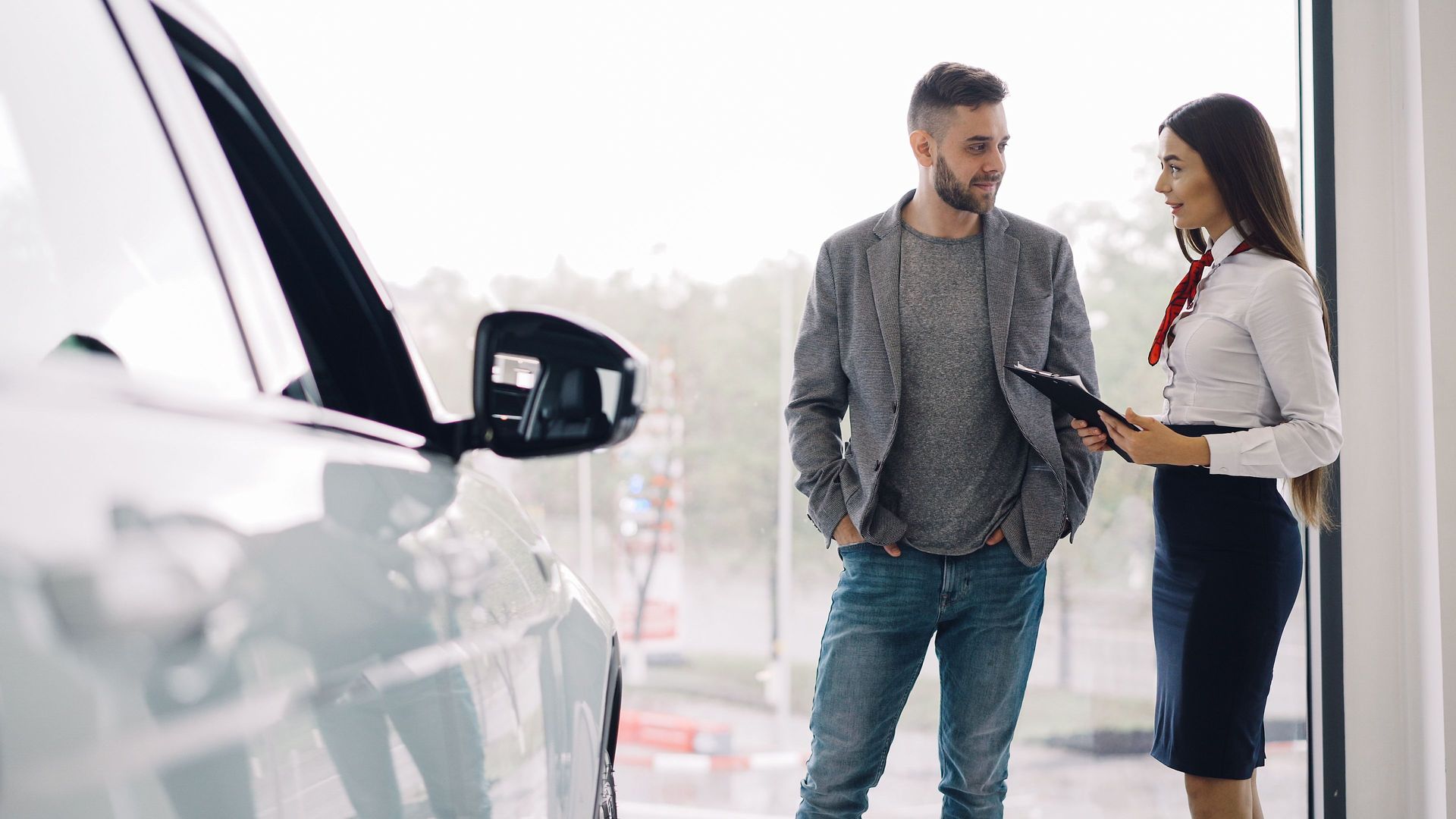 Two people talking beside a white car, with one holding a tablet in a bright outdoor setting