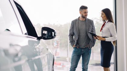 Two people talking beside a white car, with one holding a tablet in a bright outdoor setting