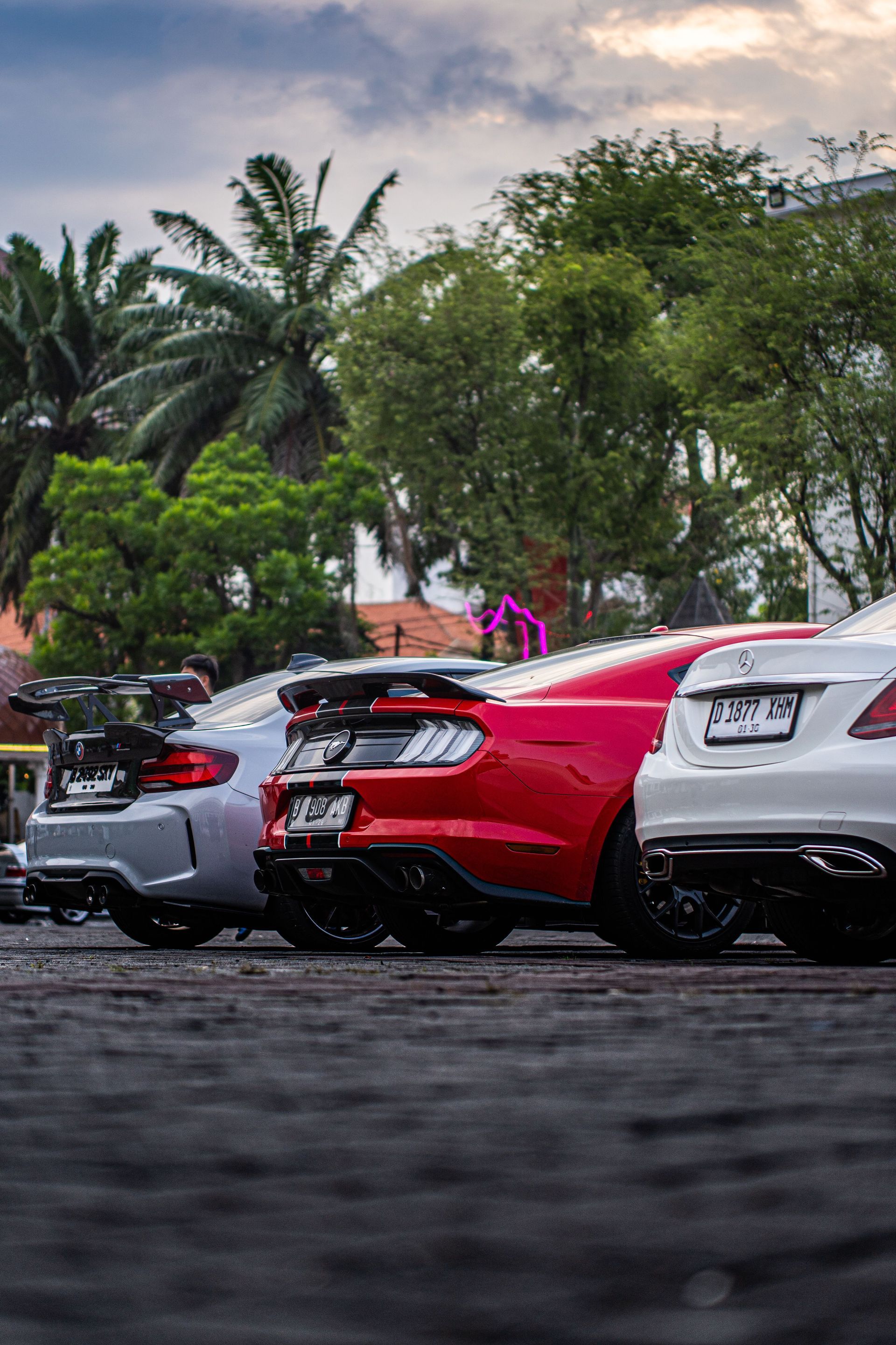 Three parked cars, including a red sports car, in a tree-lined outdoor lot at dusk