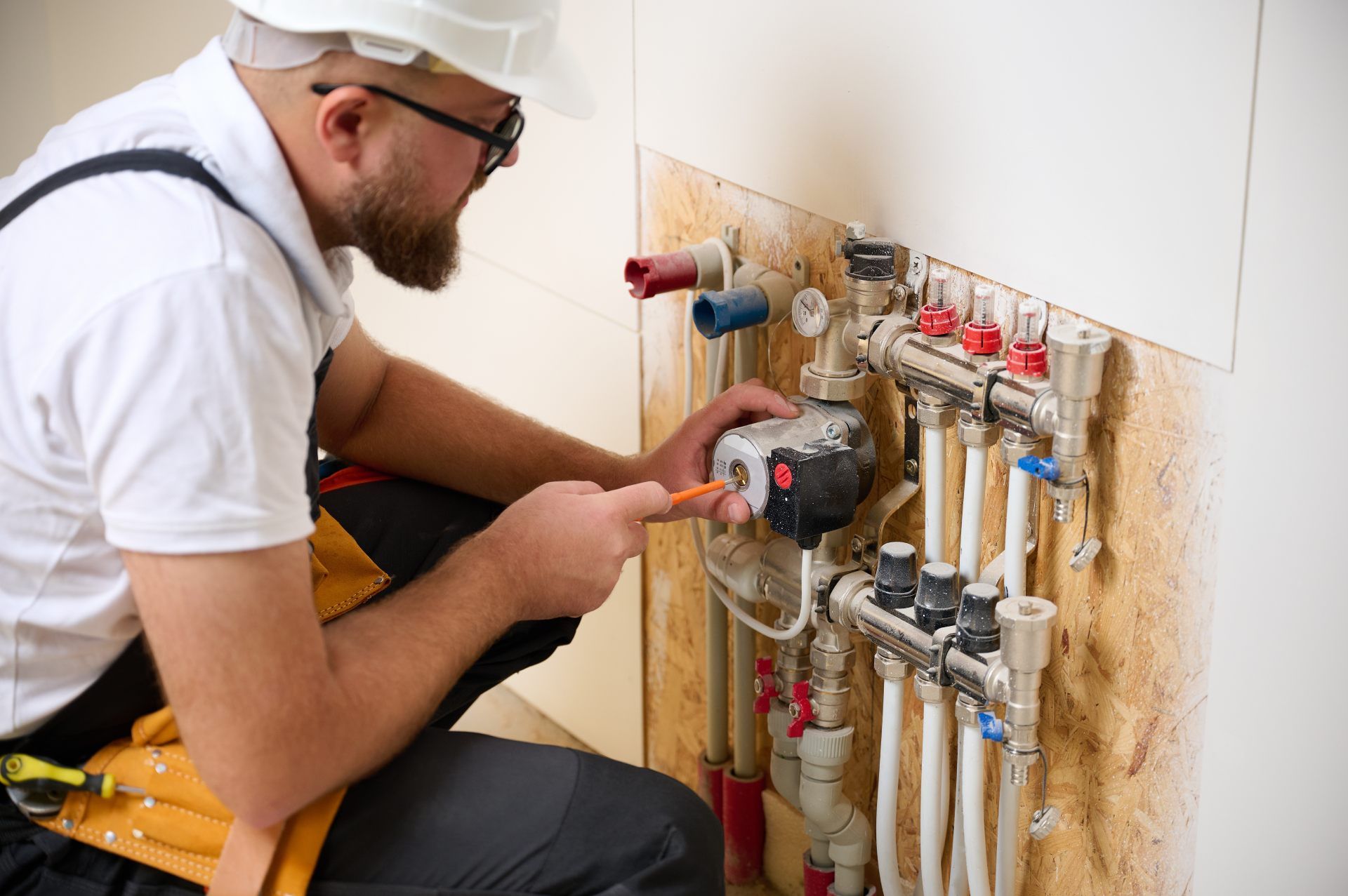 Plumber in white hard hat and safety glasses, working on plumbing pipes connected to a manifold in a wall.
