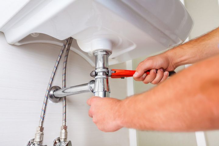 Plumber using a wrench to work on the pipes under a white sink.