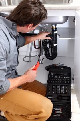 Plumber in khaki pants working under a kitchen sink with a wrench and toolbox.