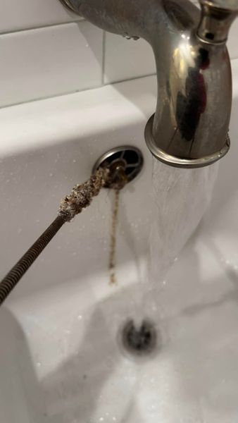 A clogged sink drain with a snake tool removing debris, water running from the faucet.
