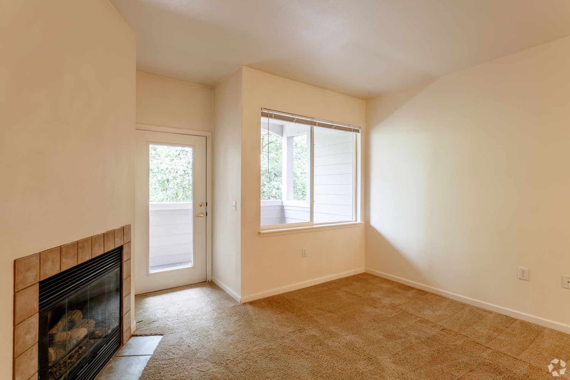 Empty beige living room with a fireplace, carpet, and windows by a balcony door.