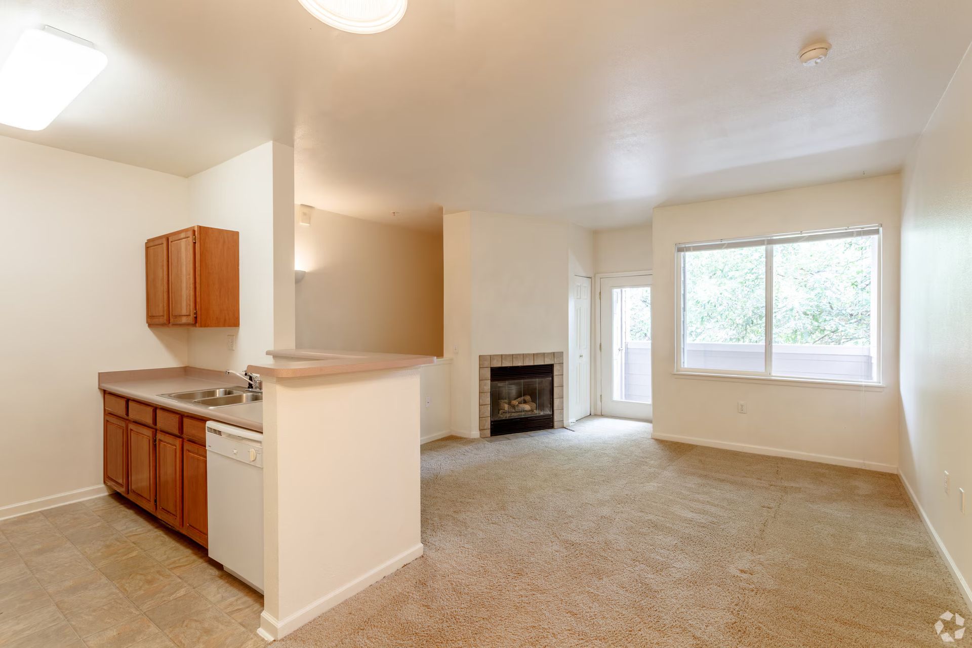 Empty beige living room with open kitchen, fireplace, and large windows