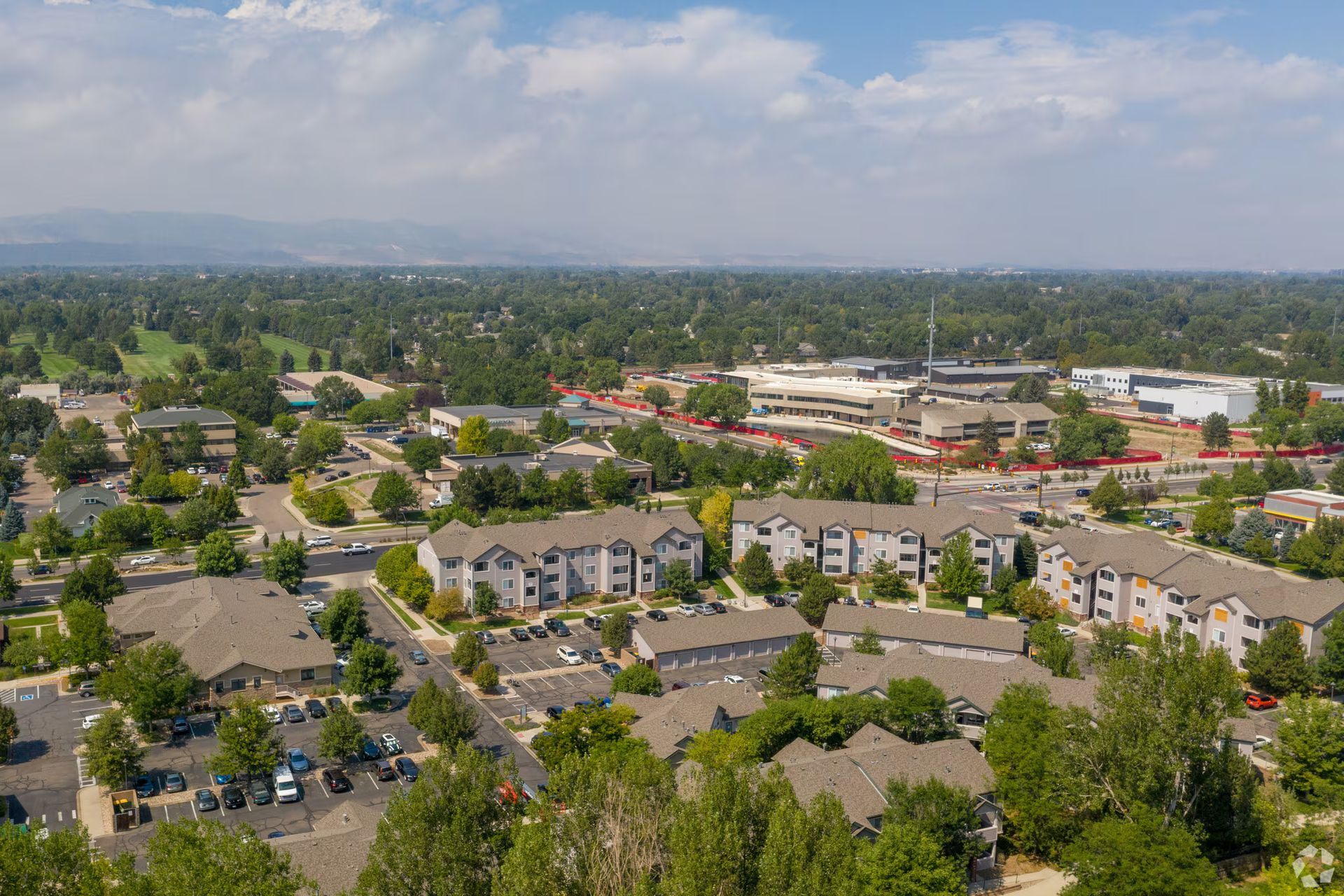 Aerial view of a suburban neighborhood with apartment buildings, trees, and distant commercial area under a cloudy sky