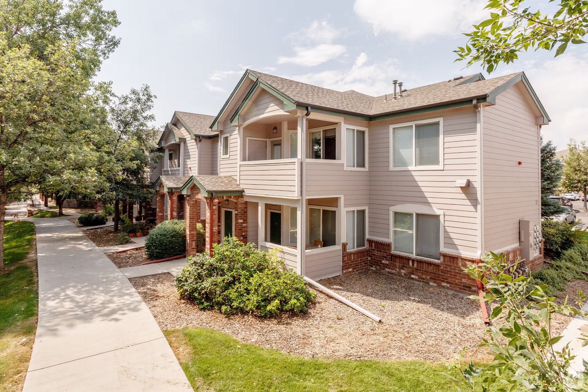 Apartment building exterior with beige siding, a sidewalk, trees, and landscaped grounds