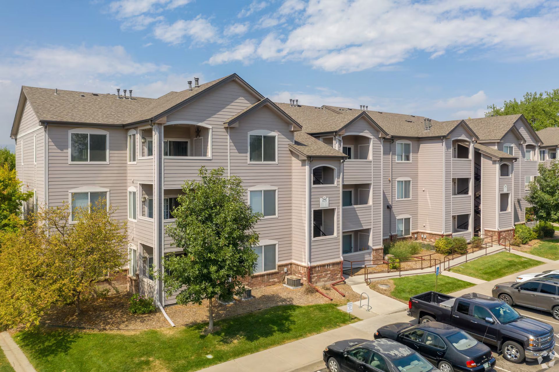 Apartment complex with beige siding, balconies, trees, and parked cars on a sunny day