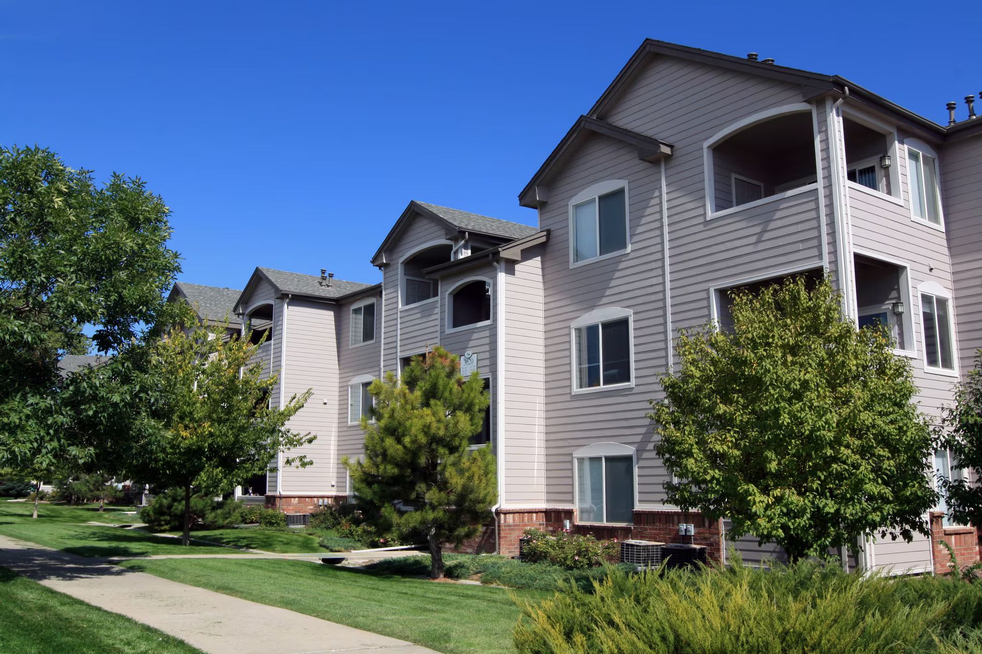 Apartment buildings with gray siding beside a sidewalk, green lawn, and trees under a clear blue sky