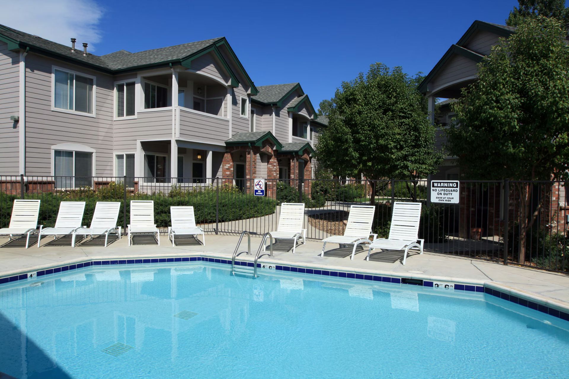 Apartment pool with white lounge chairs and tan buildings under a clear blue sky