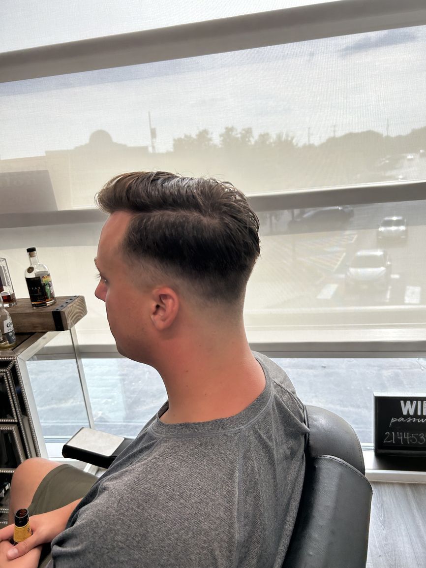 A profile view of a person with a short, faded haircut sitting in a barber chair by a large window.