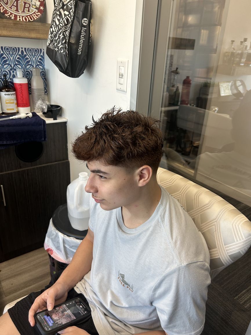 A person with textured, wavy brown hair and a faded haircut sits in a salon chair, looking at a smartphone.