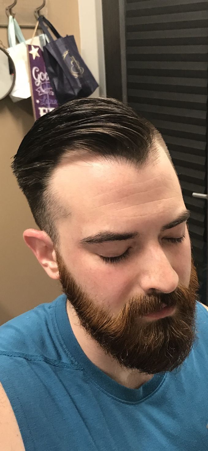 A person with a groomed beard and a short fade haircut styled back, wearing a blue tank top in a bathroom setting.