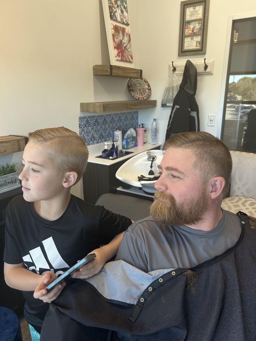 A child and an adult sitting together in a salon, both with fresh haircuts, facing the same direction.