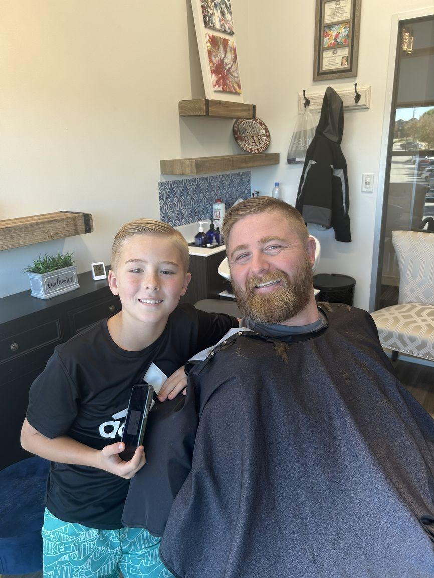 A smiling child holds a pair of hair clippers next to a smiling person wearing a barber cape in a salon setting.