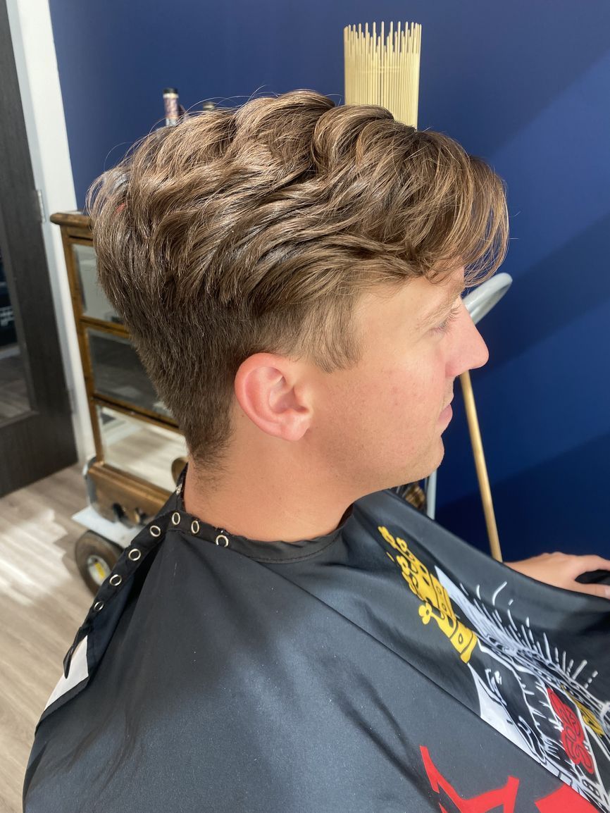 Side profile of a person in a salon cape with wavy, medium-length light brown hair and a short, faded haircut.