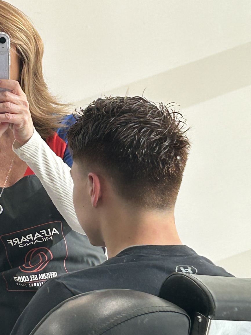 A person in a barber’s chair receiving a haircut, with the stylist holding a phone to record the process in a mirror.