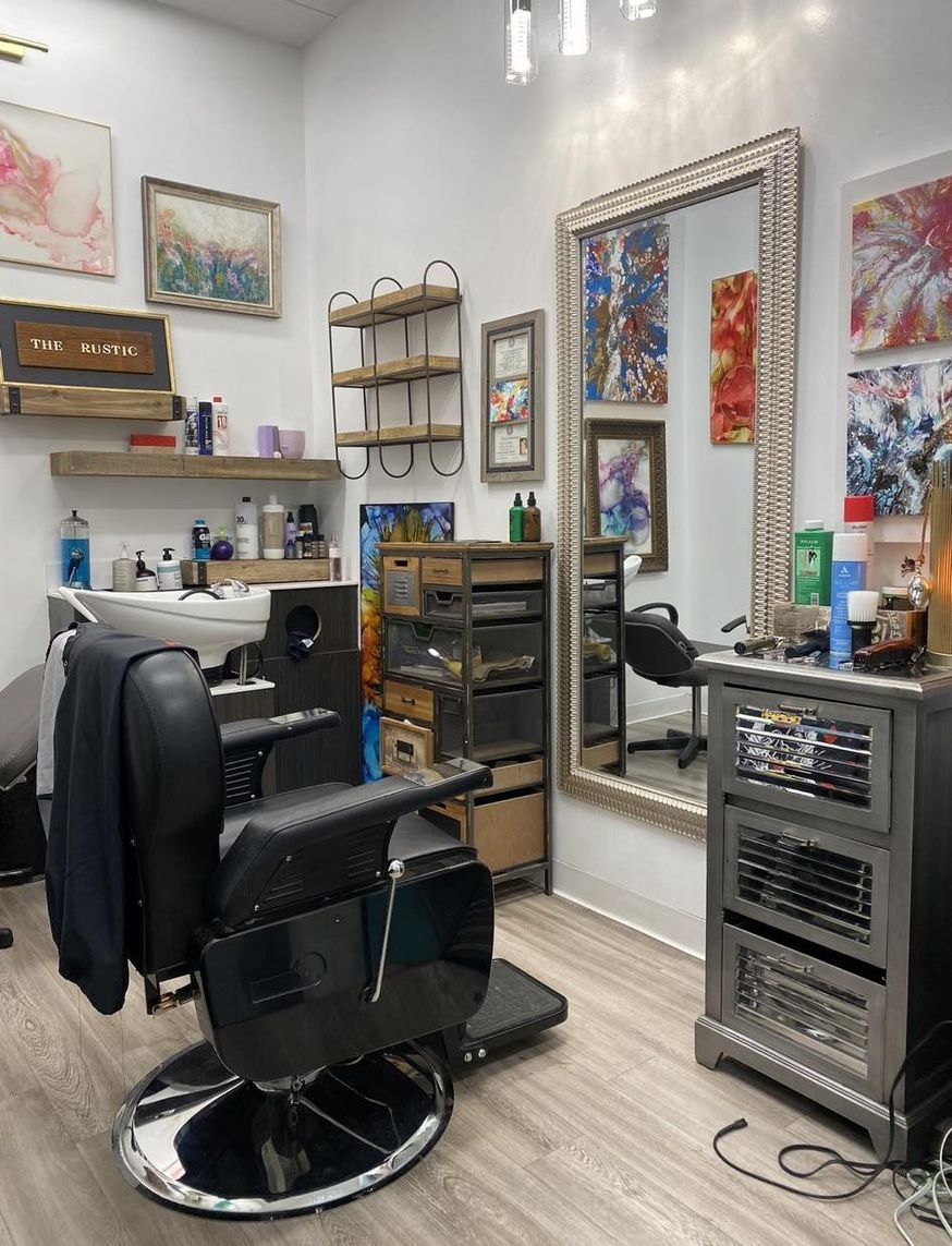 A hair salon station with a black reclining barber chair, a large mirror, a rolling cart, and wall-mounted art.