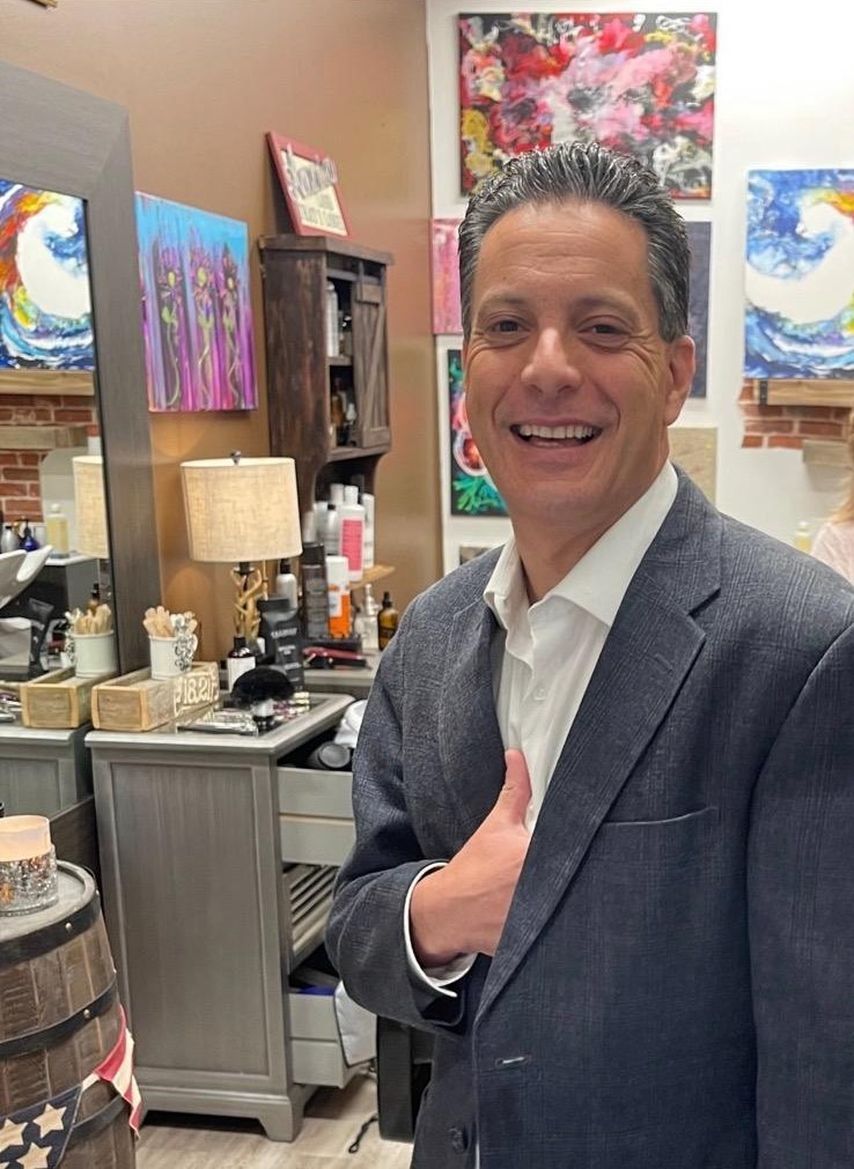 A smiling person in a blue blazer and white shirt stands in a room decorated with colorful paintings and a wooden cabinet.