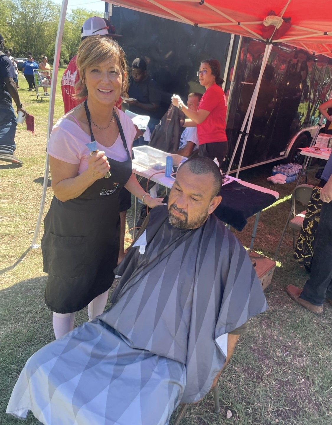 A stylist in a black apron cuts hair for a person in a cape under a red canopy tent at an outdoor event.