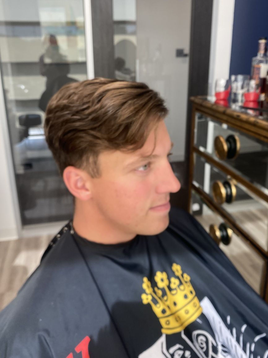 A person in a barber cape sits in a salon chair with their side profile visible, showing short, light brown wavy hair.