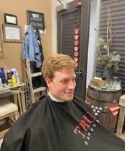 A person sits in a barber chair wearing a black cape, smiling while looking toward the right in a salon setting.
