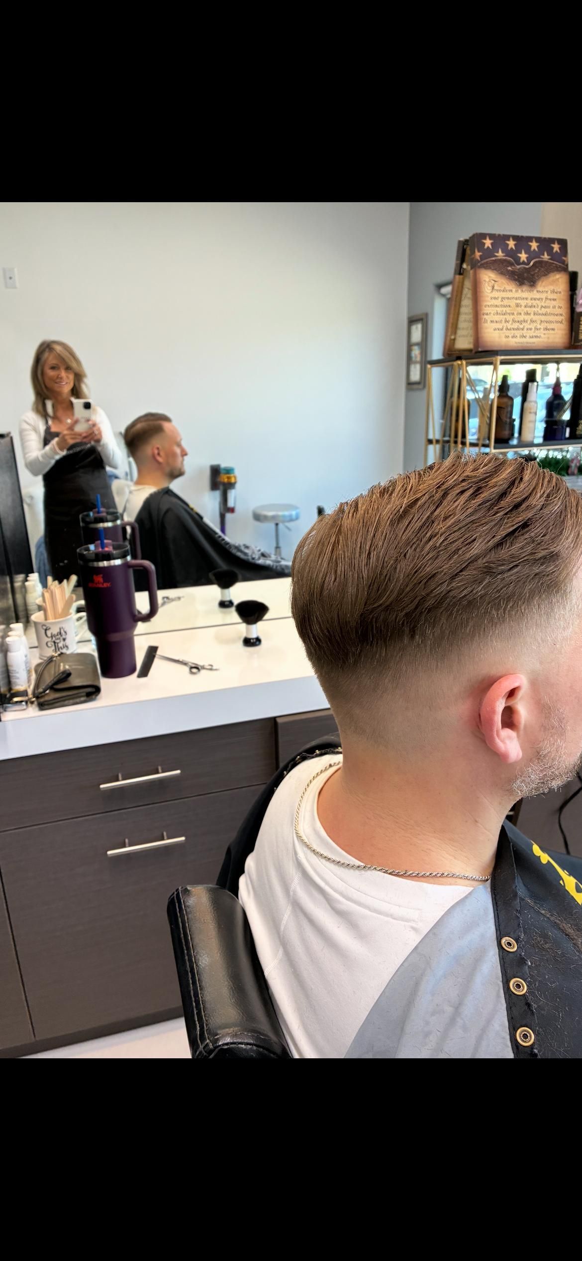 A person sits in a salon chair in front of a mirror, showing a fresh, short, faded haircut while a stylist stands nearby.