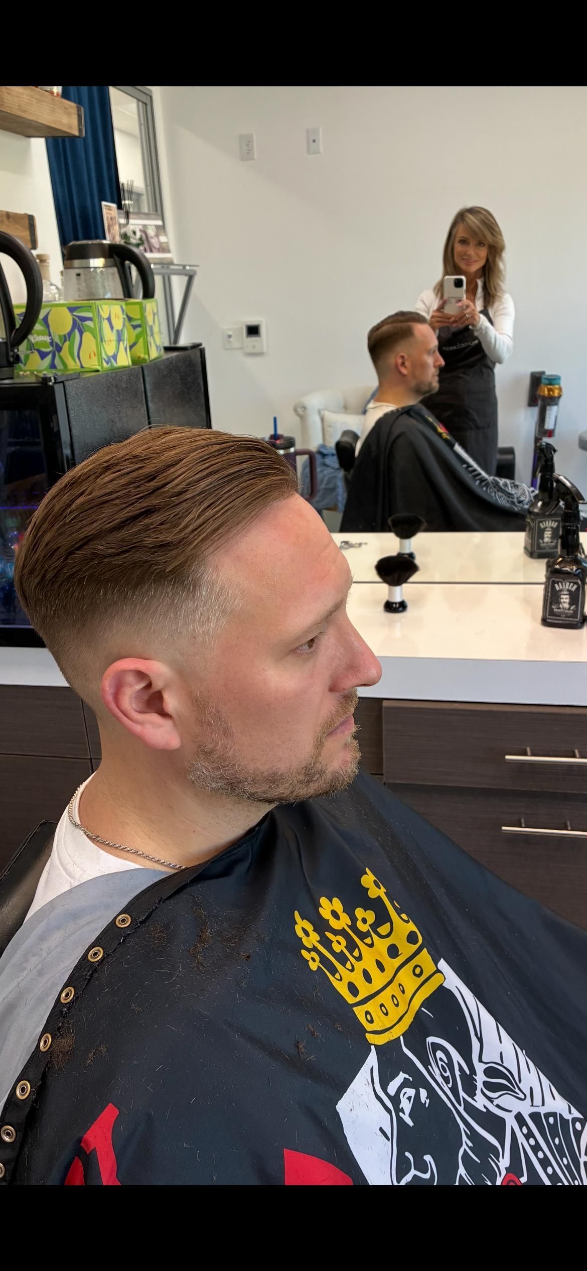 A person with a short, faded haircut and a beard sitting in a barber's chair, with the stylist visible in the mirror.