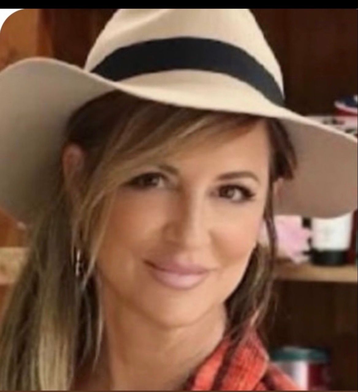 A person wearing a wide-brimmed tan hat with a dark band, posing against a soft-focus indoor background.