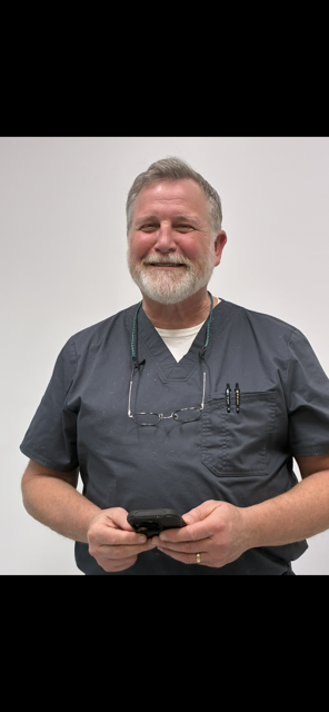 A smiling person with a beard wearing grey medical scrubs, holding a smartphone against a plain white background.