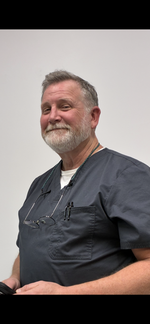 A person with a beard and gray hair, wearing a dark gray v-neck scrub top, smiles while looking toward the camera.