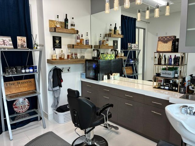 A barbershop interior featuring a black styling chair, white counter, floating wooden shelves with liquor, and a mirror.