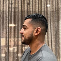 A side-profile view of a person with a short, faded haircut and a short beard, wearing a grey t-shirt indoors.