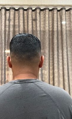 A back view of a person with a short, faded haircut wearing a grey shirt, standing in front of brown curtains.