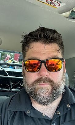 A man with a full, grey-speckled beard wearing orange-tinted sunglasses, captured in a selfie from inside a vehicle.