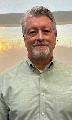A person with grey hair and a goatee, wearing a light green button-down shirt, smiles in front of a bright window.