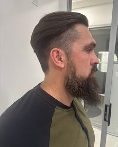 A man with a groomed beard and an undercut hairstyle seen from the side, wearing a green and black long-sleeve shirt.