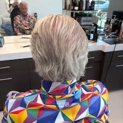 A person with silver hair sits in a salon chair, facing a mirror, wearing a vibrant, geometric-patterned shirt.