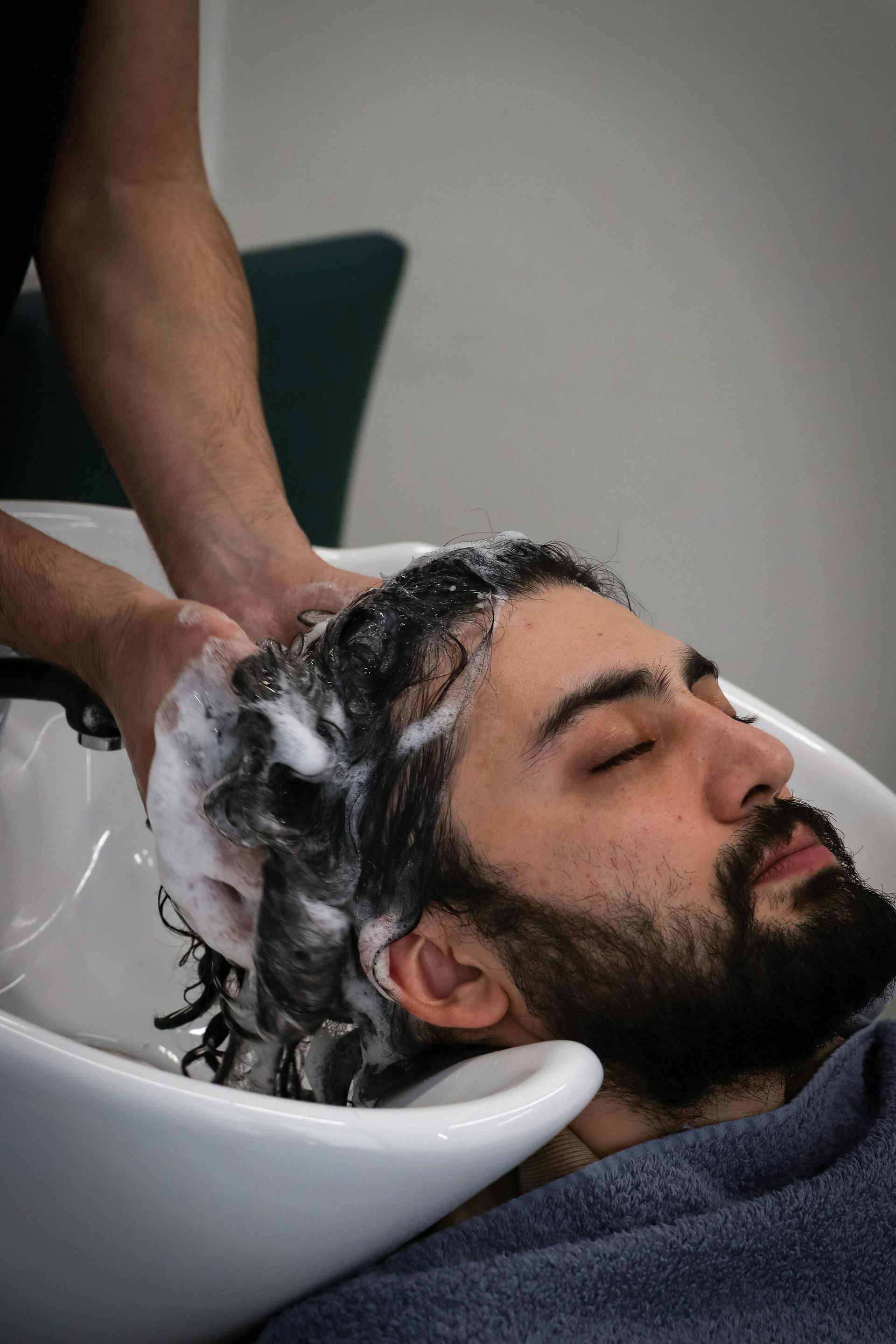 A person lies back in a salon sink with eyes closed while a stylist lathers shampoo into their dark, curly hair.