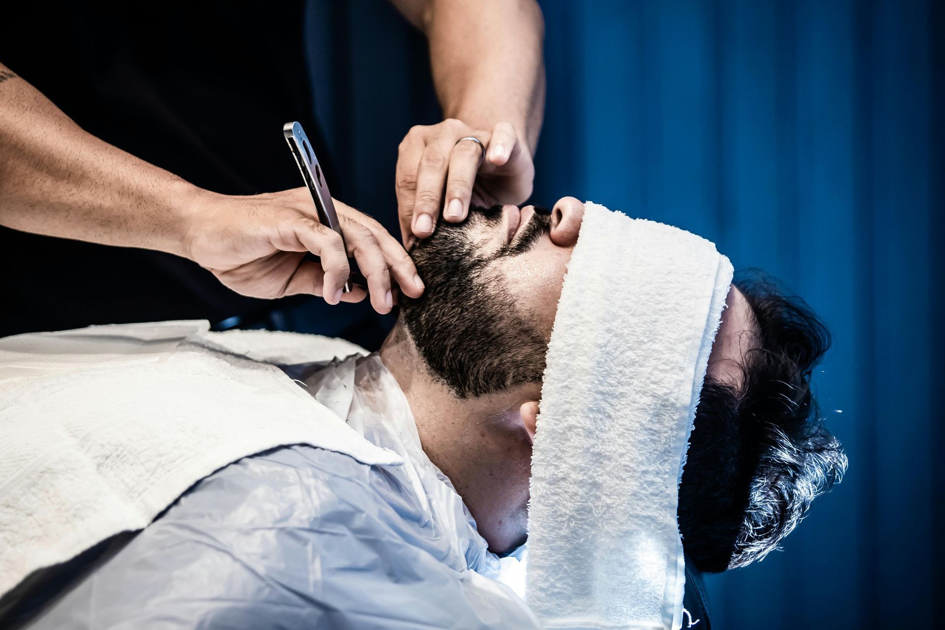 A barber uses a straight razor to groom a client's beard, with a white towel covering the client's eyes.