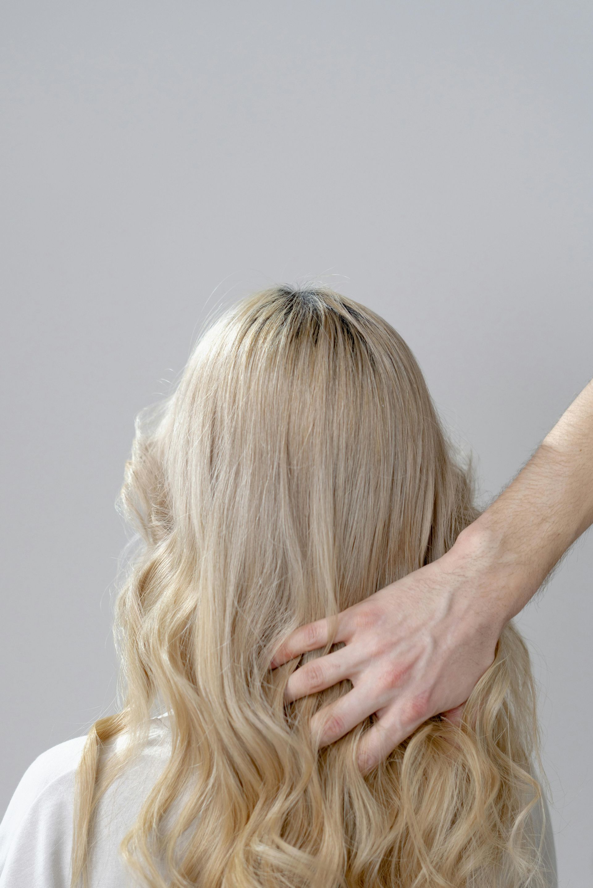 A hand gently touches the long, wavy, blonde hair of a person from behind against a plain grey background.