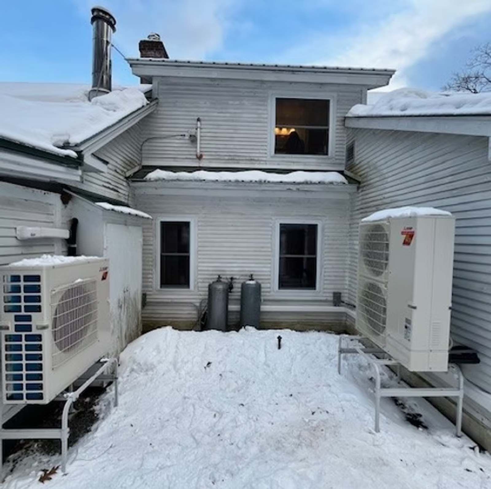 Two air conditioners are sitting outside of a house covered in snow.