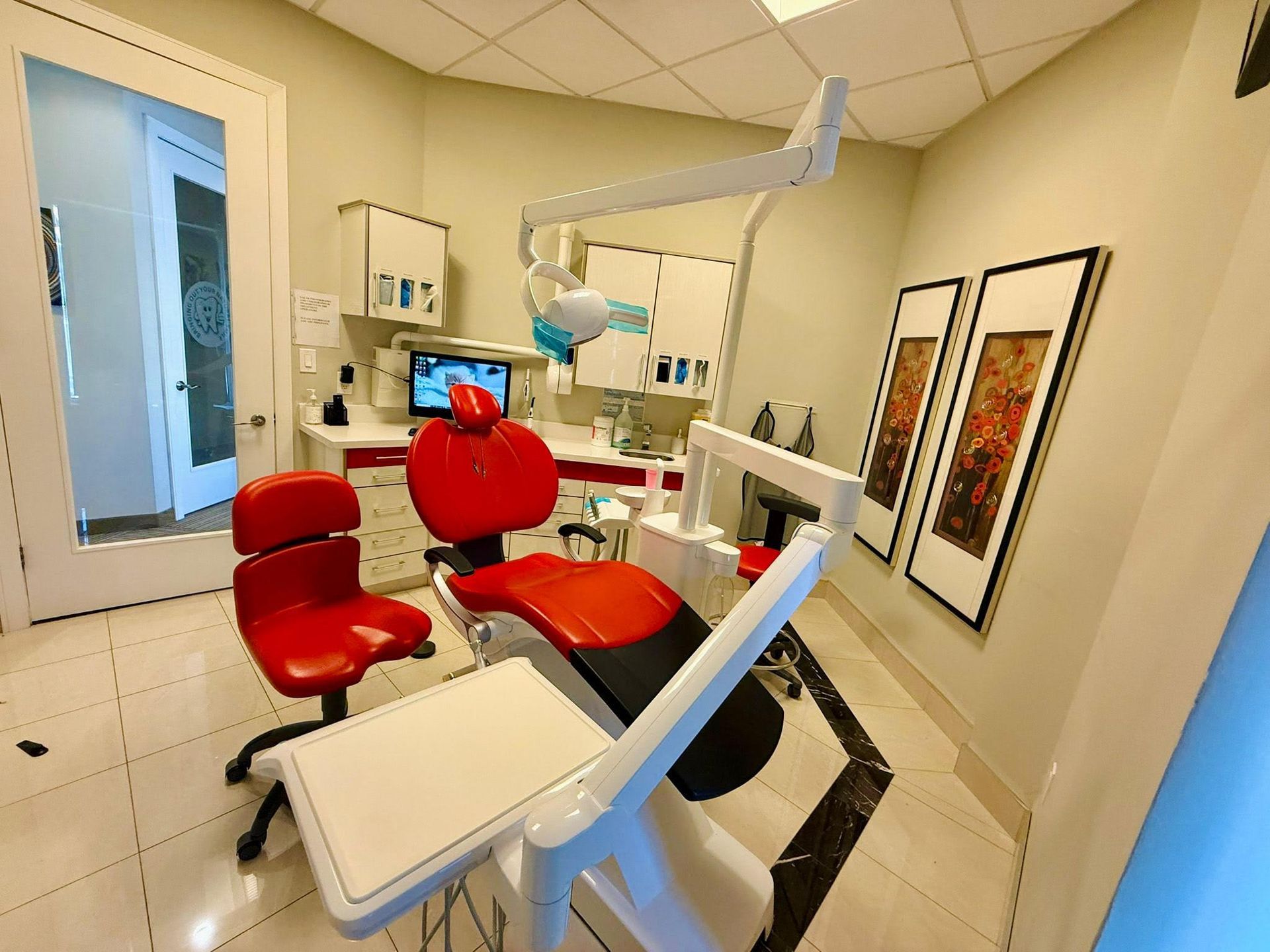 Dental operatory with red chair, white cabinets, and framed artwork.