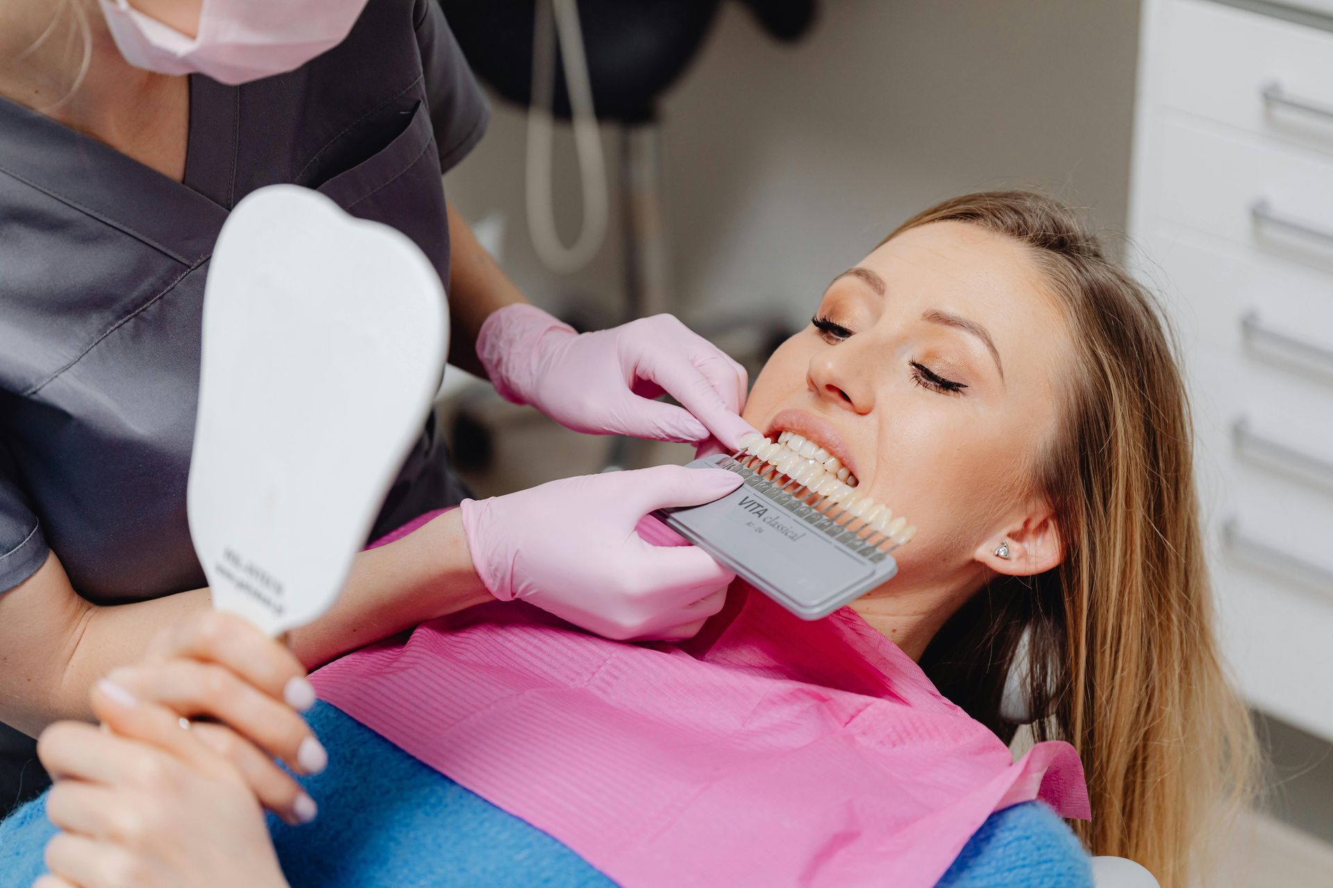 Dentist comparing tooth shade with color guide for a patient. Pink-gloved hands, dental office setting.