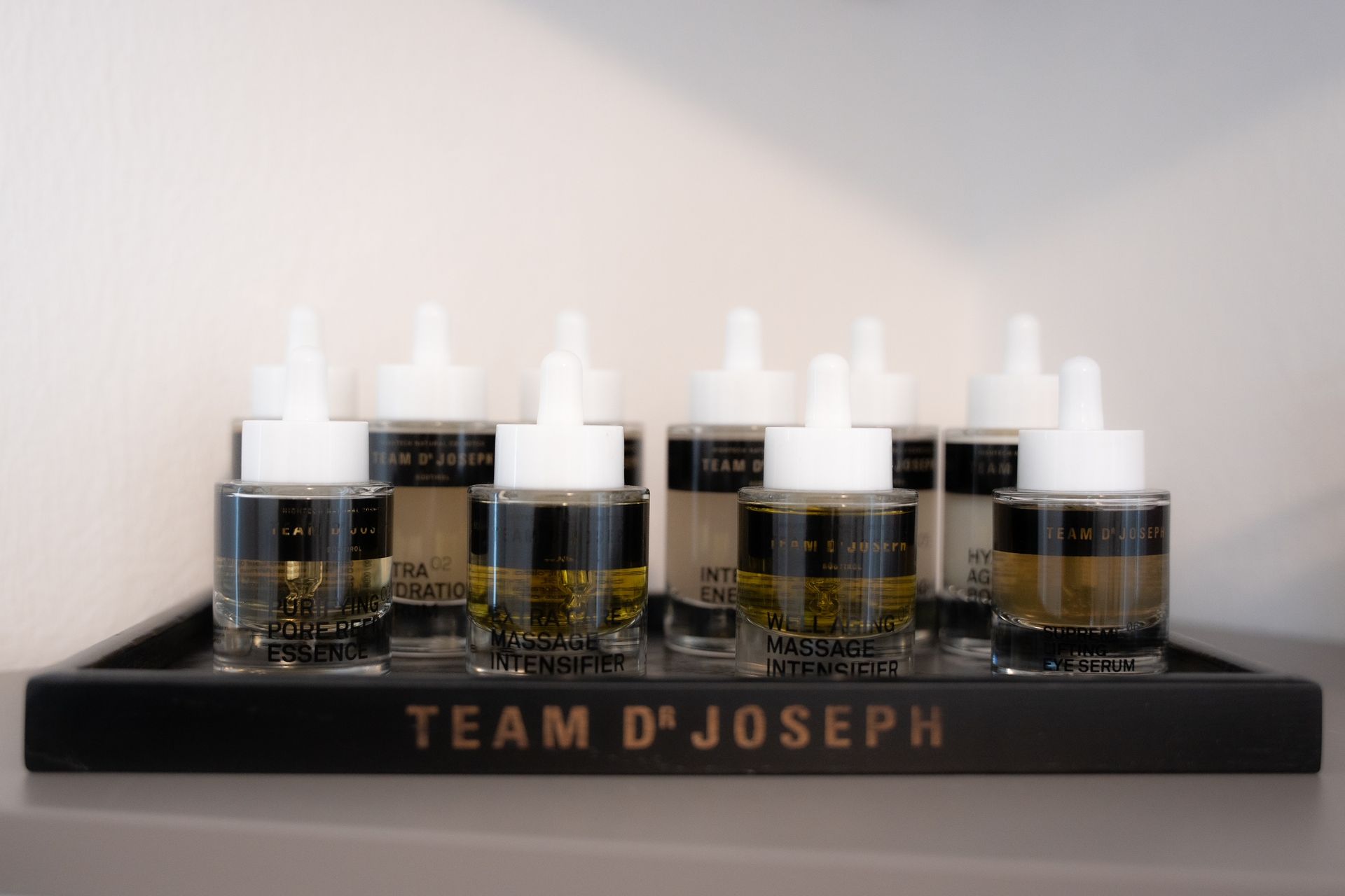 Weiße Hautpflegeprodukte-Fläschchen mit Deckel stehen auf einem schwarzen Tablett von TEAM D'JOSEPH vor einer hellen Wand.