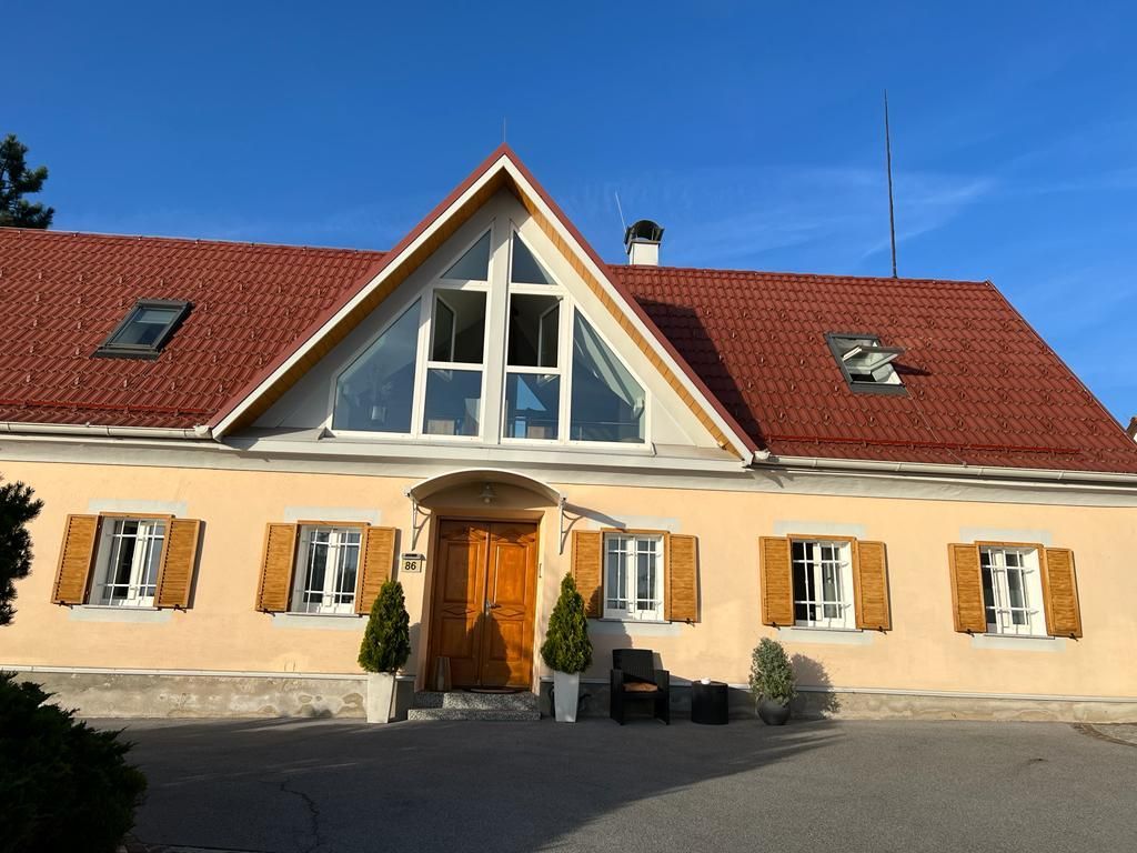 Sonnendurchflutetes, cremefarbenes Haus mit rotem Dach, Holztür und zentralem Giebel unter klarem, blauem Himmel