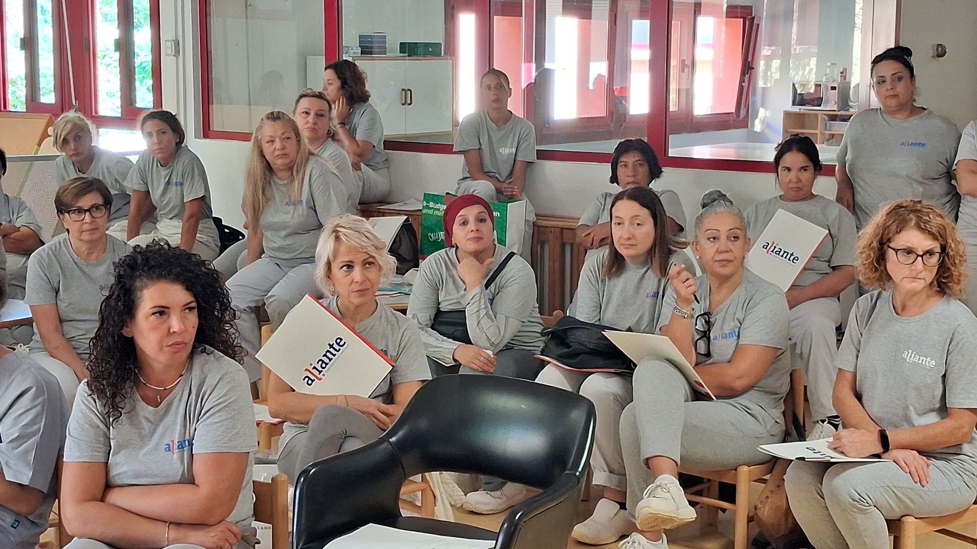 Un gruppo di persone in abiti grigio chiaro è seduto all'interno, alcune tengono in mano dei documenti.