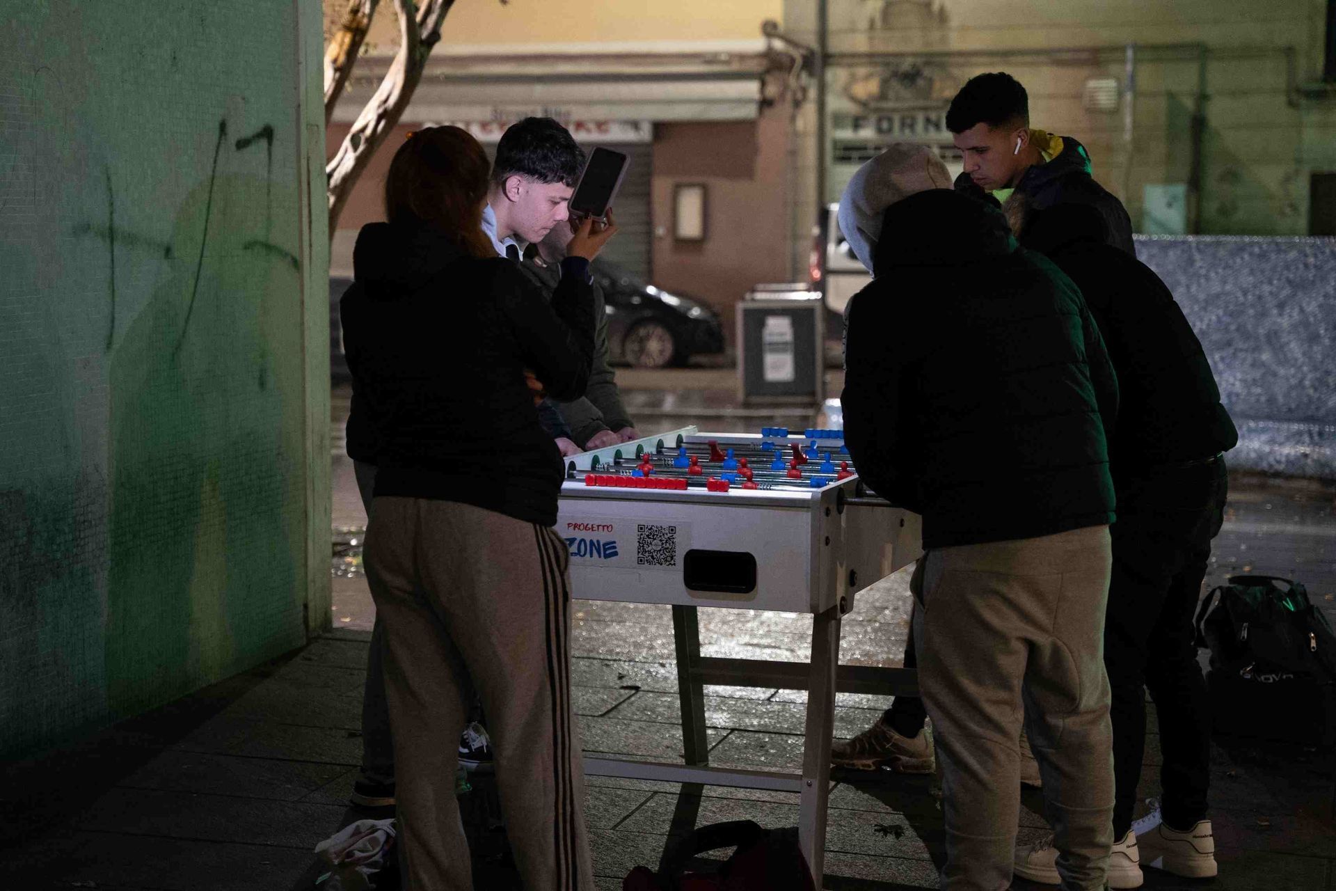 La gente gioca a calcio balilla per strada di notte. I giocatori indossano abiti casual.