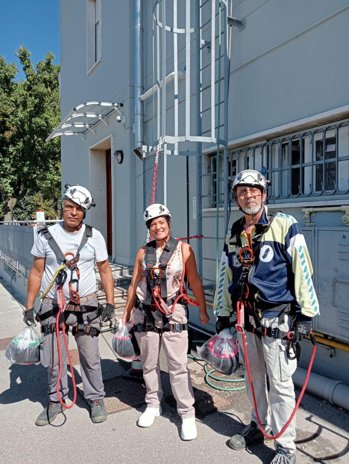 Tre persone con imbracature di sicurezza e caschi vicino a un edificio con una scala.